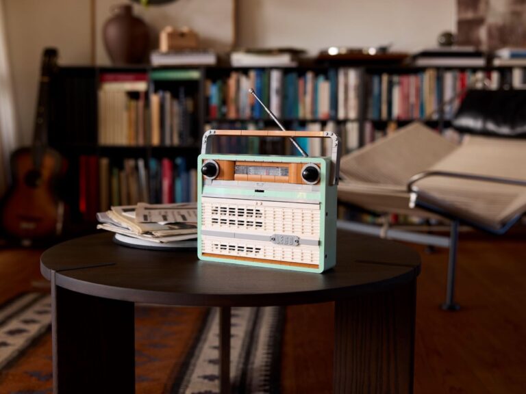 LEGO 10334 Retro Radio piparmētru zaļā, smilškrāsas un baltā krāsā ar izvilktu antenu uz tumša apaļa galdiņa dzīvojamā istabā, fonā grāmatplaukts un ģitāra