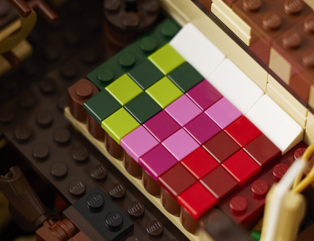 LEGO gultas tuvplāns ar pārklāju no tumši zaļām, laima, purpursarkanām, rozā, sarkanām un baltām plāksnītēm brūnā rāmī mājiņas iekšienē