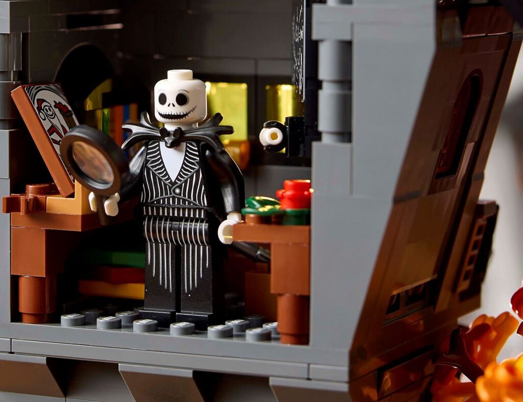 Jack Skellington minifigūra melnā svītrainā uzvalkā ar lielu palielināmo stiklu rokā, starp koka rakstāmgaldu ar atvērtu grāmatu, kurā redzams Ziemassvētku vecīša zīmējums, un galdiņu ar sarkanu un zaļu dāvanu